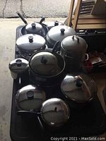 Wide view of the lot showing multiple pots and pans stacked together, mostly metal with glass or metal lids featuring black knobs, includes a multi-tiered stacking pot.