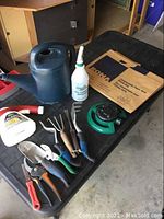 Photo showing garden hand tools, watering can, spray bottle, garden twine, green hose nozzle or sprinkler, and compostable paper yard waste bags.