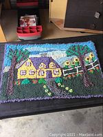 Full view of the antique hooked rug on a dark surface showing house, trees, flowers and sky.