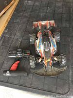 Remote control off-road car with black and orange flame decals and large tires, alongside its black and red transmitter controller on tiled floor.