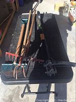 Photo shows a collection of six garden and yard tools with wooden handles placed on a black folding table.