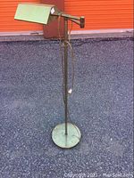 Full view of the vintage metal floor lamp with rectangular green shade and round metal base placed outdoors on asphalt in front of orange door.