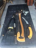 Full view of seven tools arranged on black surface including pruning shears, climbing axe, saw, wire cutters, utility knife, and lantern.