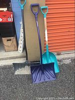 Front view of three snow shovels with different colored plastic blades and handles standing on pavement against a beige board and orange metal door