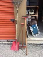 Photo showing five garden tools including a metal rake, a plastic shovel, a garden hoe, a garden rake, and an aerator with metal spikes against a tan wall next to a red door