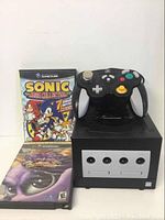 Front view of black Nintendo GameCube console with controller resting on top and two game cases standing behind.