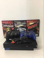 Photo showing PlayStation 2 console, two wired controllers (black and blue), three game cases (Dark Cloud, Need for Speed: Most Wanted, Burnout Revenge), and composite AV cable.