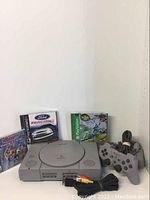 Sony PlayStation console with three game jewel cases behind it and a controller alongside, cables in front.