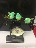 Metal clock with three green birds sitting on a metal branch with a clock hanging beneath.