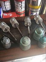 Top-down view of six glass insulators, four mounted on metal rods with flange bases, two loose bluish-green dome-shaped insulators, displayed on wooden surface with fire extinguishers in background.