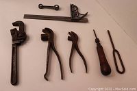 Photo shows a heavy duty adjustable wrench, two pairs of pliers, a wood handled screw starter, and a metal tool with loop handle arranged on a white surface.