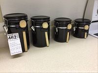 Full view of the four black ceramic canisters in descending order of size, each with a sealing lid and wooden spoon attached on the side.