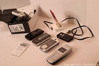 Photo showing six cell phones including a Sony Ericsson flip phone, Alcatel flip phone, and other assorted phones with chargers and packaging in the background.