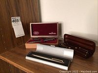 Three sets of pens and pencils displayed on a wooden shelf including a Sheaffer set in a red box, a dark wood case with burgundy pens, and a silver and black modern case.