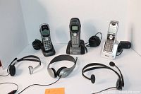 Five V-Tech cordless phone handsets with accompanying power cords and four wired headsets, showing models including a silver V-Tech phone and Plantronics wired headset.