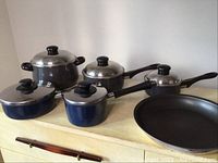 Photo of six pots with lids and one frying pan arranged on a kitchen counter, black and blue pots with black handles and knobs on lids.