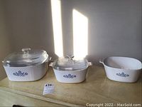 Three white Corningware casserole dishes with blue flower motif, two have glass lids, displayed on wooden surface with sunlight casting shadows on wall.