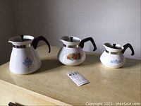 Front view of the three Corningware tea pots in varying sizes with lids on a wooden surface.