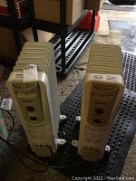 Two upright beige oil-filled electric heaters by Noma positioned side by side on a black mat, shown from front perspective, measuring 11x14x25 inches.