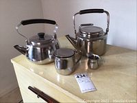 Set of two stainless steel kettles, small creamer, and tea steeping pot on yellow table against white wall.