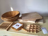 Photo showing two handmade wooden salad bowls stacked, three wooden cutting boards in the background, and three wooden trivets arranged in front including a slatted trivet, a grid trivet, and a tile-handled round trivet.