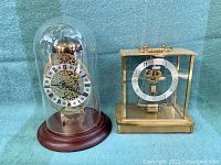 Two vintage European style clocks, one with glass dome and wooden base, the other with brass frame and glass sides, shown on a blue cloth background.