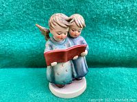 Front view of the vintage ceramic figurine showing two angelic children wearing blue gowns singing from a large red songbook.