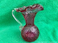 Photo showing cranberry glass pitcher with clear handle and ruffled top edge against a green background.
