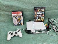 White Sony PlayStation 2 slim console with two game boxes and AV cables connected, shown on green background.