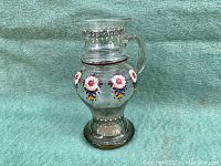 Side view of the vintage hand-blown glass pitcher showing floral enamel decoration, handle, and flared rim with swirl designs.