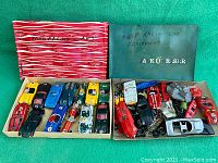 Photo of the full lot showing two open boxes with various vintage slot cars and numerous parts alongside them. Left box contains painted slot cars in racing styles with driver figures. Right box contains partially assembled bodies, chassis, and mechanical components.