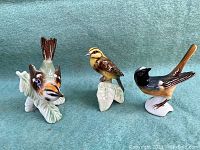 Three bird figurines on yellow-green background showing front and side views. Orange-black bird diving, yellow-brown bird on a leaf, black and brown bird on white base.
