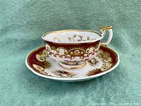 Side view of Minton cup on matching scalloped saucer showing intricate hand painted gold scroll and floral decorations with burgundy border.