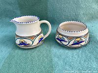 Photo showing set of two vintage pottery items: a creamer pitcher with spout and handle, and an open bowl. Both pieces are white with colorful art deco pattern including blue, yellow and orange, and rim scalloped with blue dots.