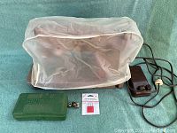 Vintage Singer sewing machine covered in clear plastic cover with foot pedal and cords to the right, green plastic case labeled SINGER in front, metal bobbins and red-packaged machine needles to the side.