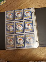 Photo showing binder page with 9 Pokemon cards placed in plastic sleeves. The cards are facing down, showing the back side with the Pokeball design.