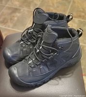 Two black KEEN men's hiking boots, size 13, placed side by side on a brown surface, showcasing their profile, laces, soles, and design details.