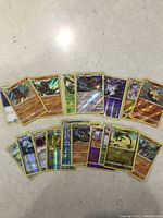 Top view of assorted 22 Pokémon trading cards laid out in two rows on a white surface, showing holographic foil and various Pokémon characters.