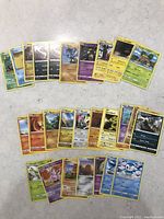 A spread of approximately 23 assorted Pokemon cards on a white surface, showing various Pokemon characters and types, arranged fanned out for display.
