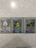 Photo shows three Japanese Pokémon holographic cards in protective sleeves laid out on a surface.