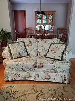 Front view of ivory loveseat with floral upholstery and two matching pillows, setting in a dining room area.