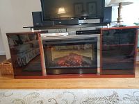 Full view of wood and glass TV stand with central electric fireplace and remote on floor in front.