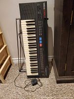 Full view of Yamaha PSR-7 keyboard set on metal stand with power adapter on floor.