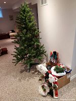 Full view of 6-foot artificial Christmas tree standing next to boxes containing various Christmas decorations including Santa figurine, wreaths, and garlands.