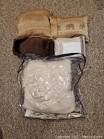 Multiple decorative pillows arranged on carpet including beige pillows with patterns and dark brown one, plastic bag contains beige bedding items