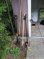 Photo of shovel, hoe, green metal fan rake, and hand saw with wooden handle placed against a shed wall and plants.