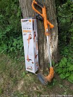 Photo showing the orange and black Worx cordless trimmer/edger tool leaning against a tree near its original box.