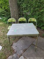 Photo of the square metal card table with three matching folding chairs arranged behind, on outdoor ground with trees in background.