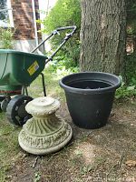 Shows Scotts Turf Builder fertilizer spreader alongside black plastic pot and concrete pedestal, outside by tree and brick wall.