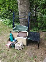 Camping gear spread out near a tree outdoors showing cooler boxes, folding cot, side tables, bags, and bottles